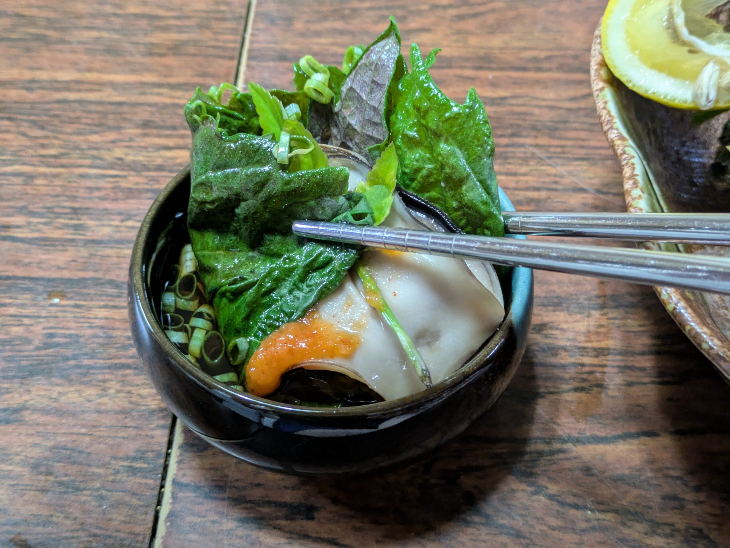 酒蒸しにした大きな岩ガキを大葉でくるみ、たくさんの薬味をつけて箸で掴んでポン酢につけている