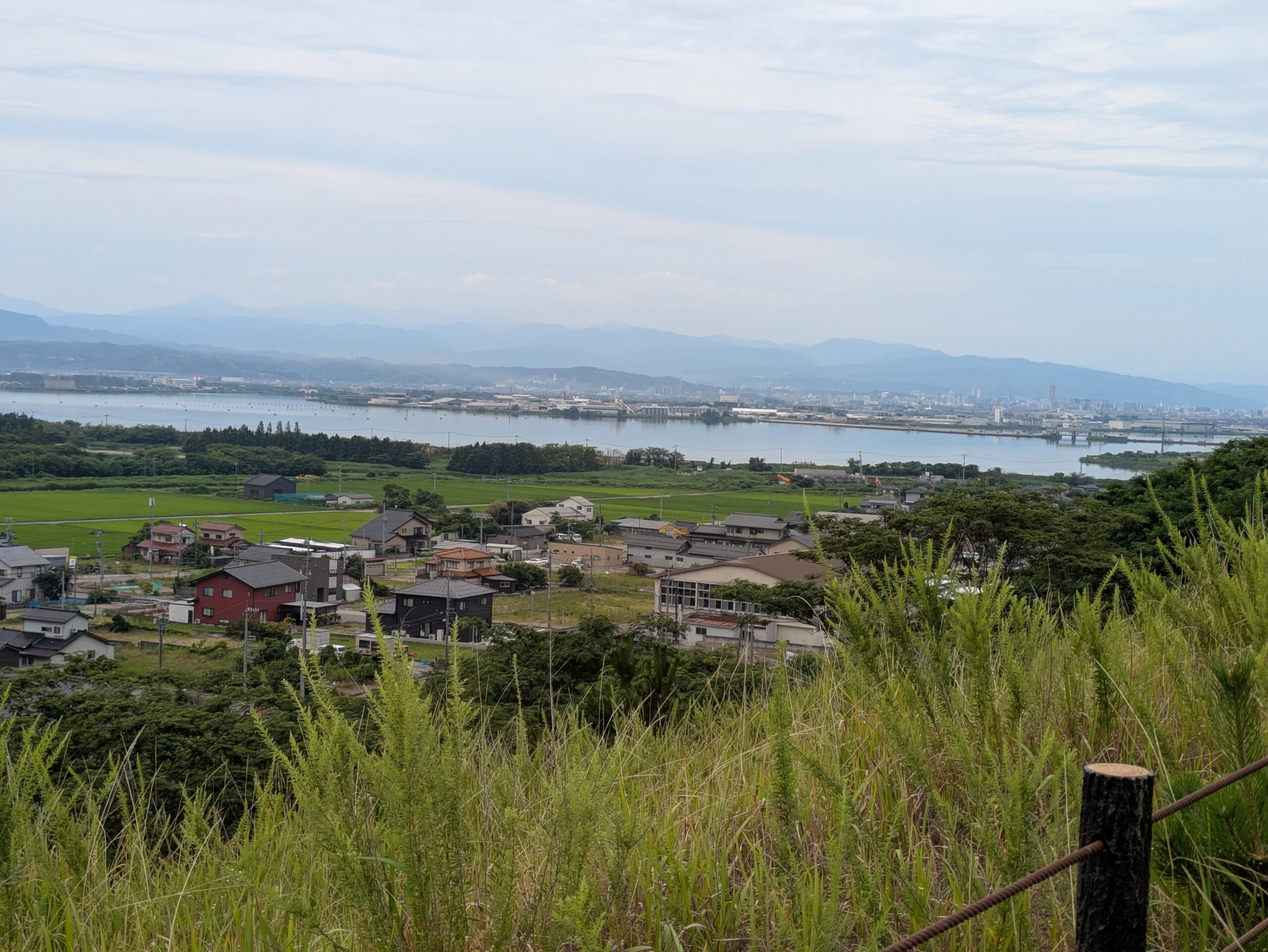 内灘砂丘の上の草むらから見下ろした景色。手前に集落や田畑が広がり、その奥に横たわる河北潟と、遠くに広がる山々が見える。