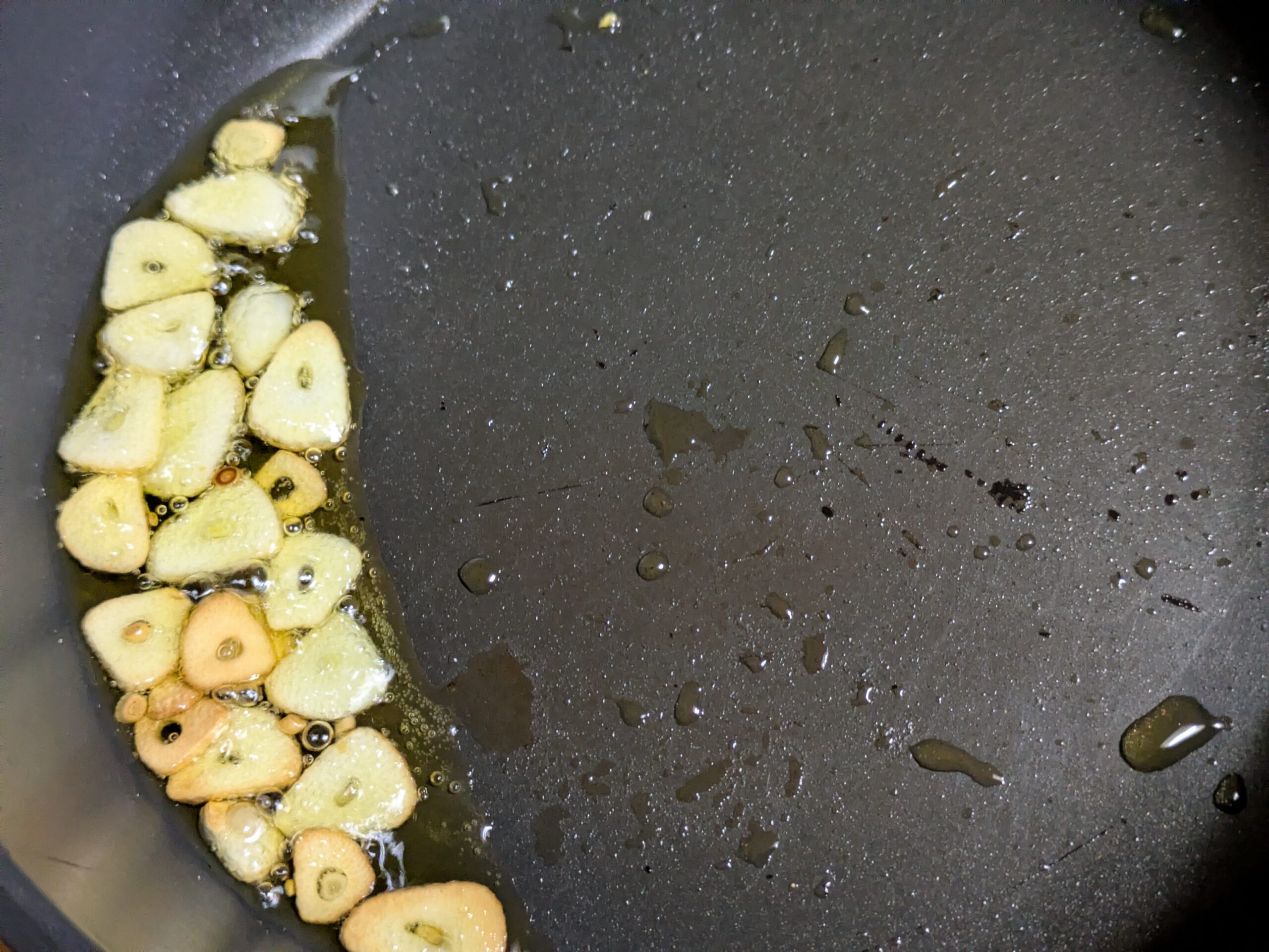 フライパンのオリーブオイルで薄切りニンニクを揚げ焼きにしている様子