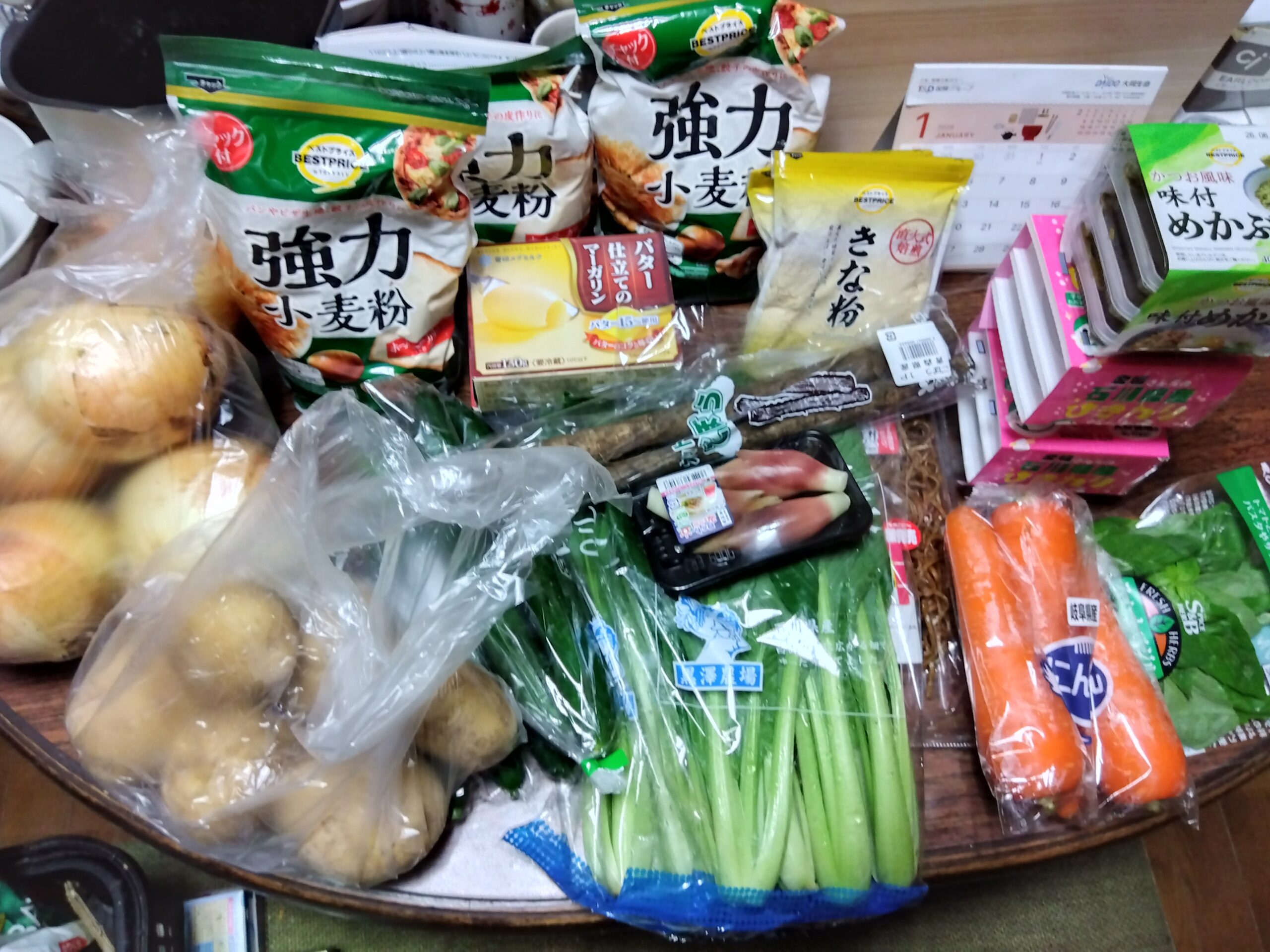 小麦粉、じゃがいも、玉ねぎ、人参、小松菜など大量の食材を机に並べた写真